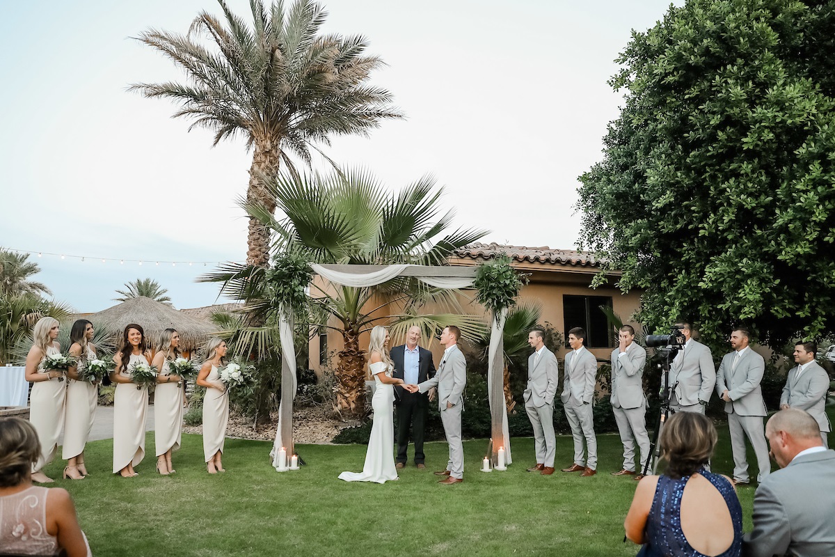 couple holding hands getting married by the officiant at the altar surrounded by the bridal party on either sides in an outdoor wedding ceremony setup